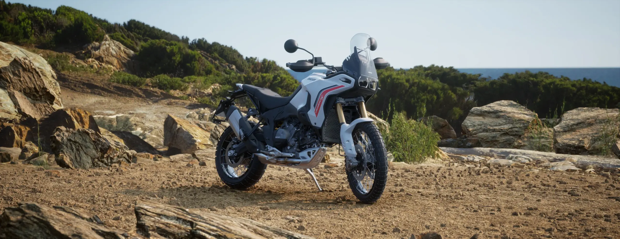 2026 Ducati DesertX white motorcycle in a front three-quarter off-road view with rugged terrain and distant mountains.