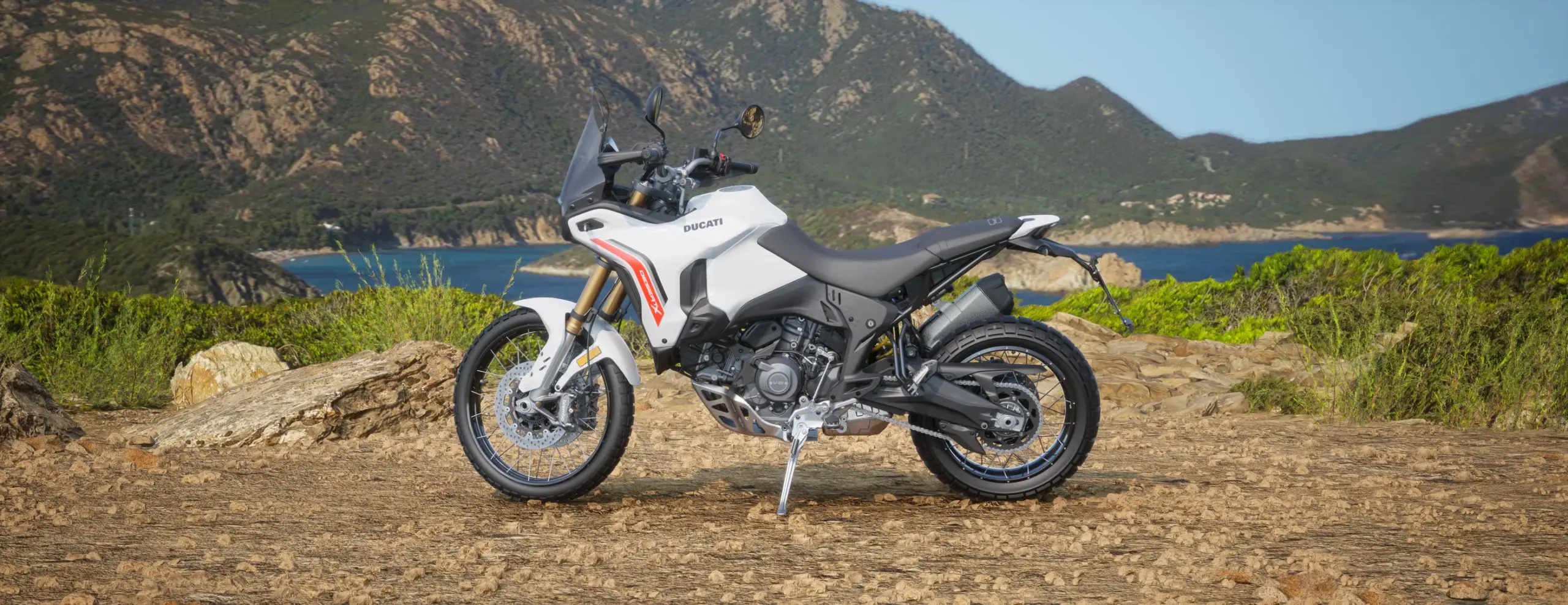 2026 Ducati DesertX white motorcycle in a left-side profile within a rocky off-road setting.